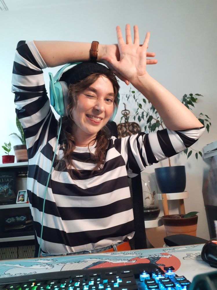 Selfie von Merri in einem schwarz/weiß gestreiften Pullover, einer schwarzen Wollmütze und Headset. Sie sitzt an ihrem Schreibtisch und zeigt mit der Hand alle 5 Finger