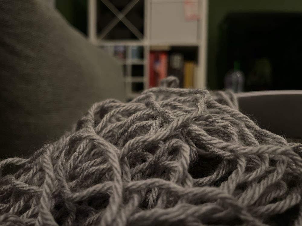 Pile of grey wool, unravelled upon a grey couch. White bookshelf in the background.