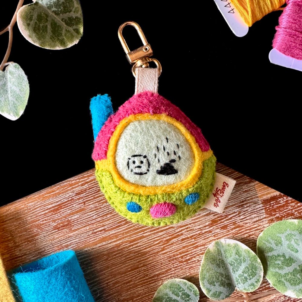 A product picture of a felt tamagotchi-like toys. It is egg-shaped, pink and green with embroidered buttons and a blue antenna. The creature embroidered on the screen is a sad blob next to a stinky poop. A ribbon linked to a golden lobster claw is attached to the top of it.