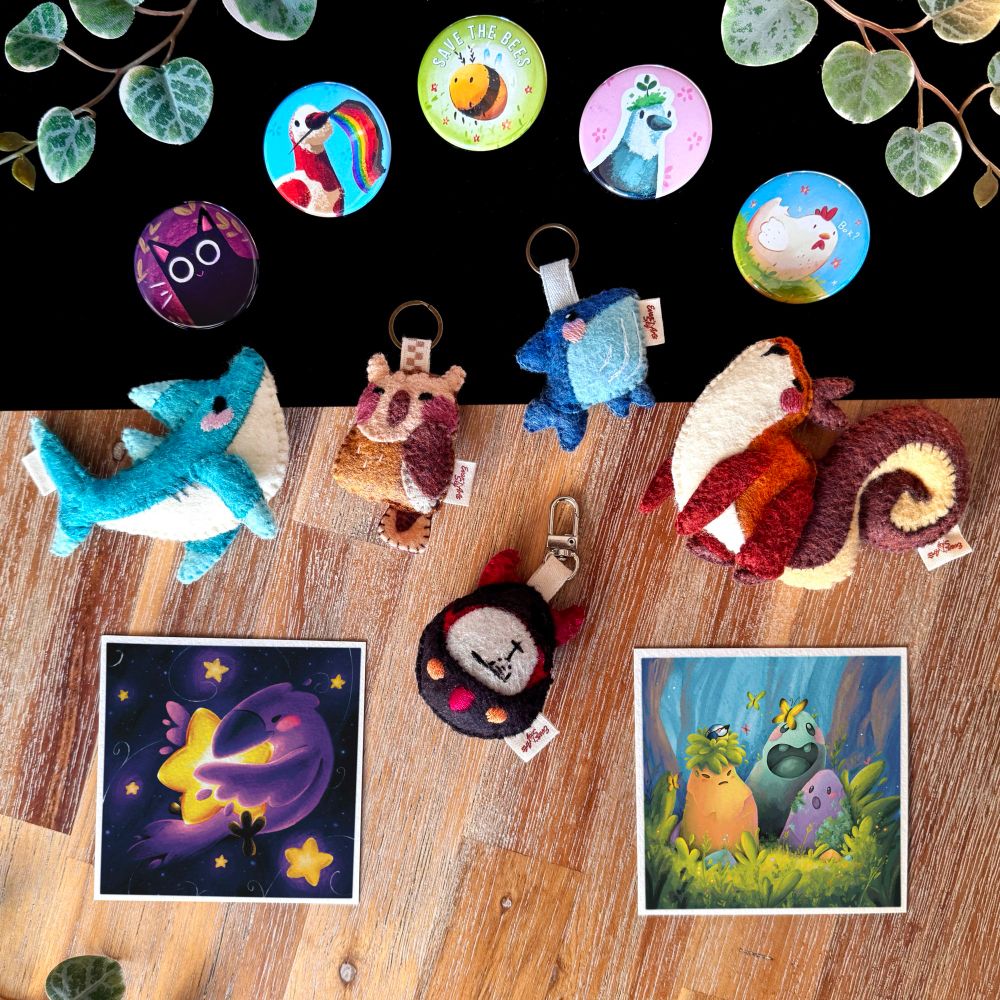 Picture of several products on a black and wooden background, framed with some decorative plants. 
On top are 5 button badges: a black cat, a brown and white pigeon holding a pride flag, a bee with the words « save the bees » written, a grey and white pigeon with a patch of grass on its head, representing the aromantic flag; and a white hen on a patch of grass. 
Underneath, a shark plushie, an owl keychain, a whale keychain, a squirrel plushie, and a black tamagotchi with a tombstone on the screen; all handmade with felt.
At the bottom, two square prints, one with a Raven floating in space and hugging a big star, and another one with three magical stones hanging out on the forest. 