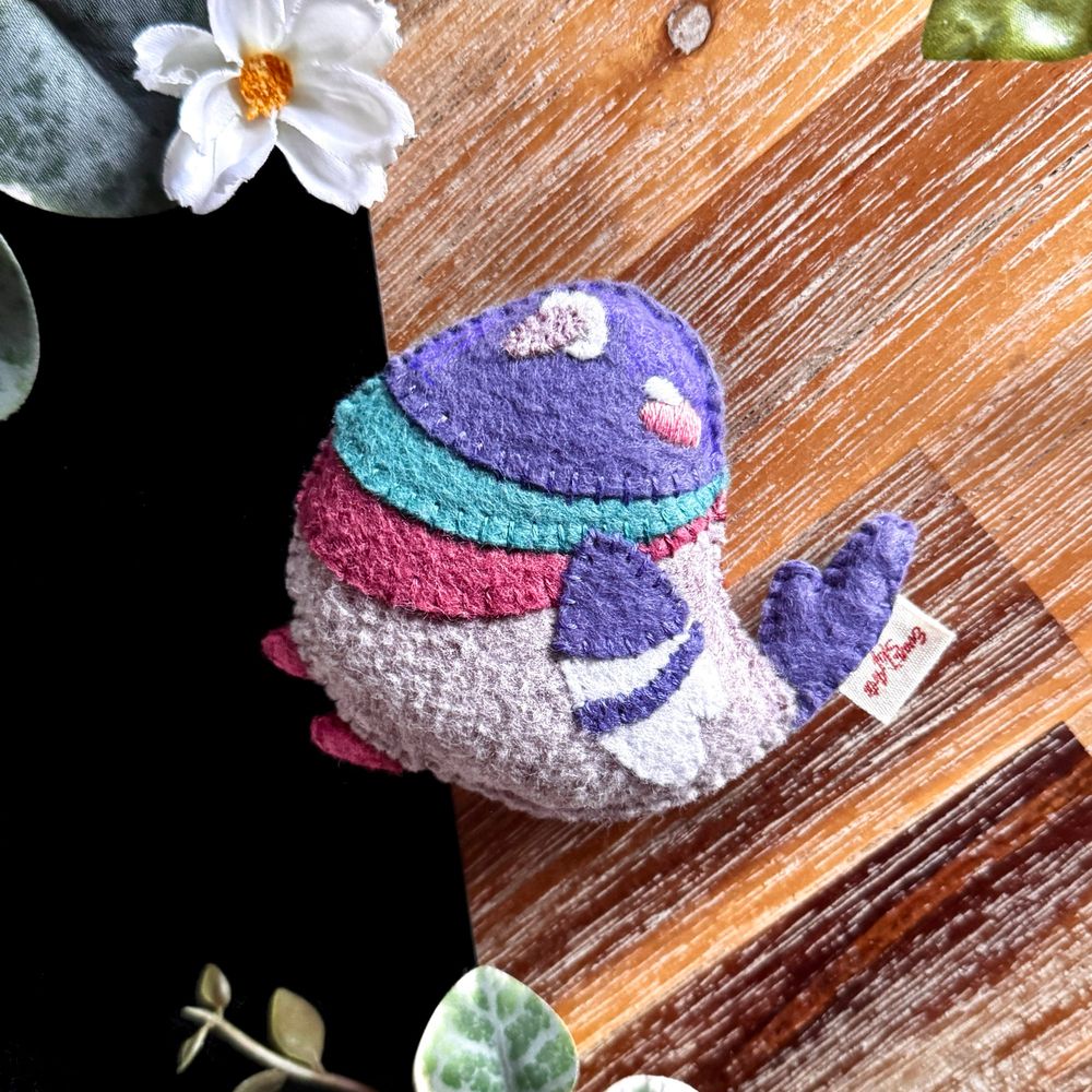 A picture of a small handmade felt pigeon plushie. Its body and beak are a warm grey with a purple head, wings and tail. It has two stripes on its neck: one teal and one a dark vintage pink that matches its two little feet. Its nose is white, as well as the two stripes on its wings. It has a happy and satisfied look on it’s face, with white embroidered closed eyes and a little pink blush.