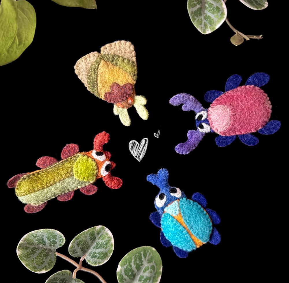 Four handmade felt magnet gathered around on a black background, a few decorative plants surrounding them and two little white heart doodles in the center. 
There are three beetles (a pink, a blue and a long green one) and a small green and brown moth. 