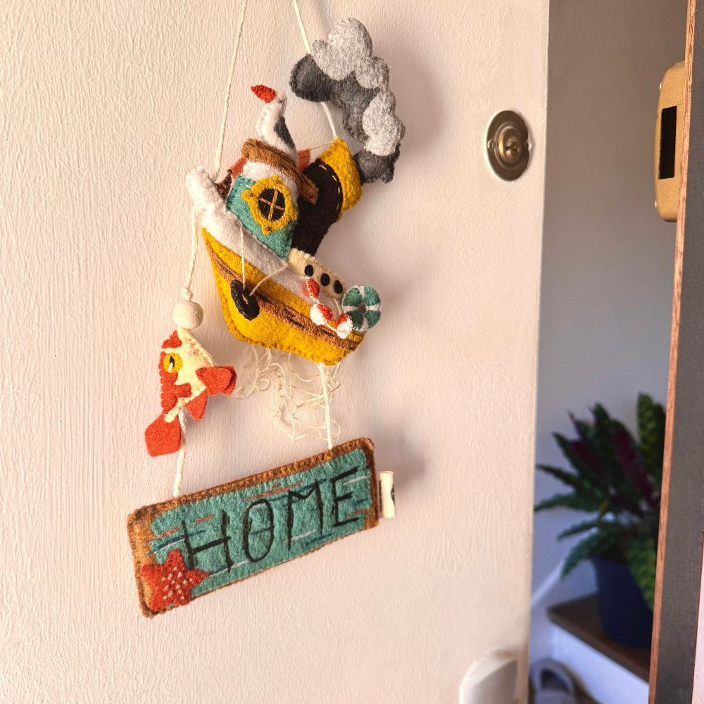 On a felt sign hanging on a slightly open front door.  The sign feature a white, yellow and blue fishing boat with a lot of little features such as hanging floaters and a big, black and yellow chimney spitting out thick grey smoke.  A seagull is perched on the roof and a fish is hanging off one of the threads that links the boat to a felt rectangle sign that reads “HOME”, the words embroidered on what looks like blue paint chipping off a wooden board. All in felt.