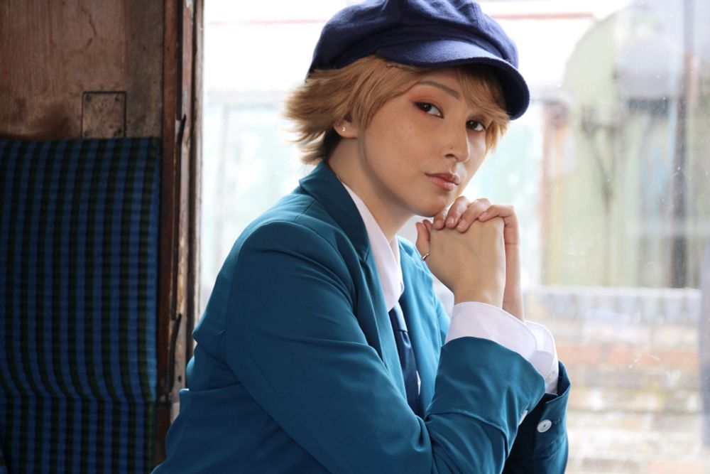 a portrait photo of andy taken on a steam train carriage in clive cosplay. they are resting their chin on their clasped hands and looking pensively at the camera 