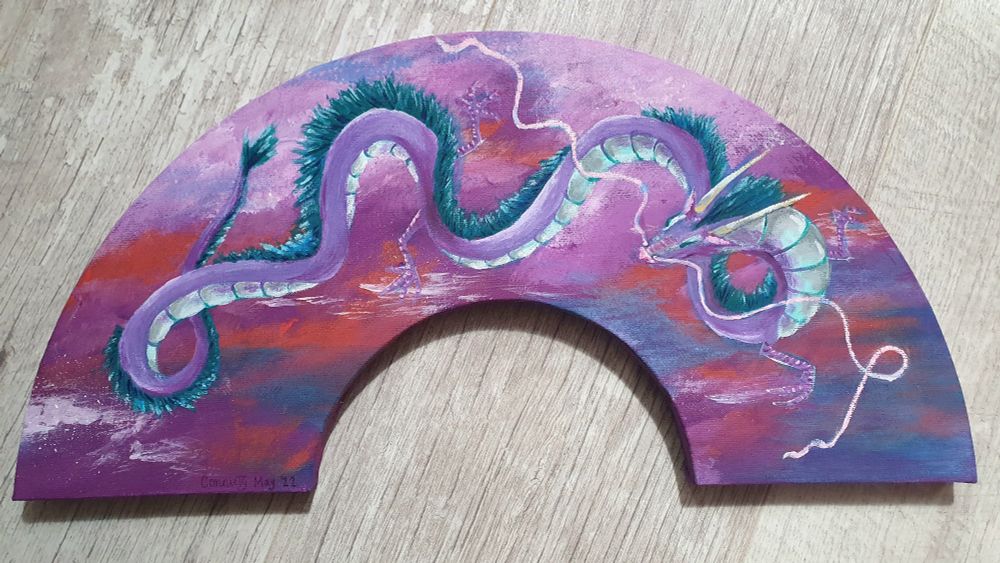Photo of an acrylic painting on an arch shaped canvas board. The painting depicts a lilac and teal dragon, inspired by Haku from Spirited Away. The main body of the long spiralling dragon is lilac, with a white underbelly. The dragon has a long mane of teal fur with a fluffy tail. It has long white horns and long spiralling tendrils coming from its snout. The background is a deep sky of dark blue, red, pink and purple with some fluffy clouds.