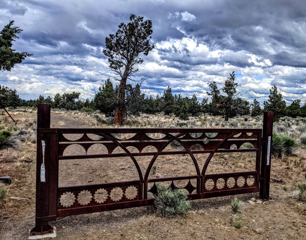 Ornate iron gate at the trailhead of high desert road into the Bandlands. Dry grass and junipers dot the landscape. Dark clouds in the sky.