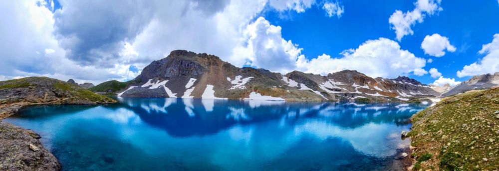 Blue lake water reflecting mountains with a little snow. Blue sky with clouds above.