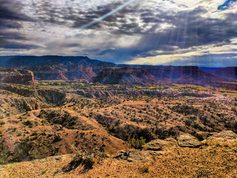 Storm clouds with rays of sun above red flat top mountains with gold trees and town below. Trail traverses hilltop of red desert.