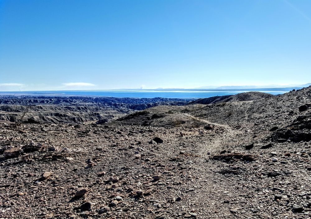 Just a lonely trail in black gray dirt, rocks, and sand leading to mountains and canyons. The Saltón Sea is in the background. 