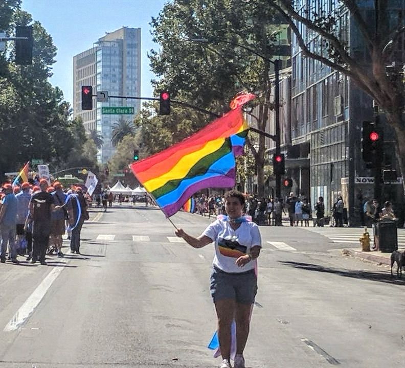 People marching for #pride down Market Street in San Jose, CA. Woman breaks out from crowd and runs down the street with rainbow flag waving behind her. 