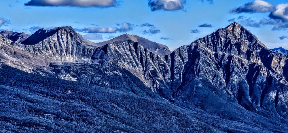 Gray mountains with blue sky with gray puffy clouds. Gray shaded trees in foreground. 