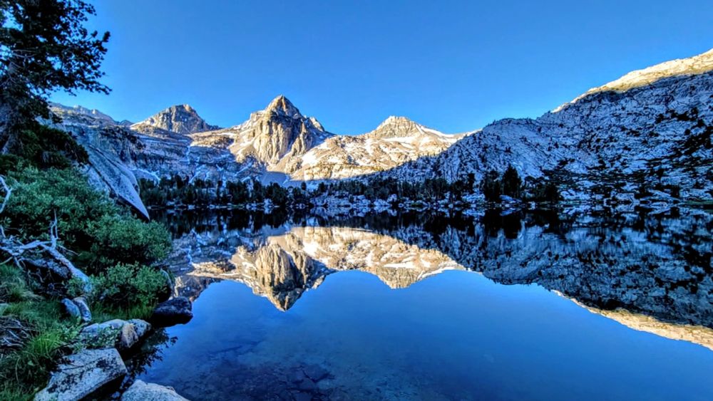 Sunlit mountain peaks with clear blue sky reflected in calm lake.