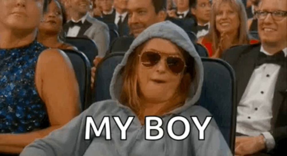 a woman wearing a hoodie and sunglasses is sitting in a crowd of people and says `` my boy '' .