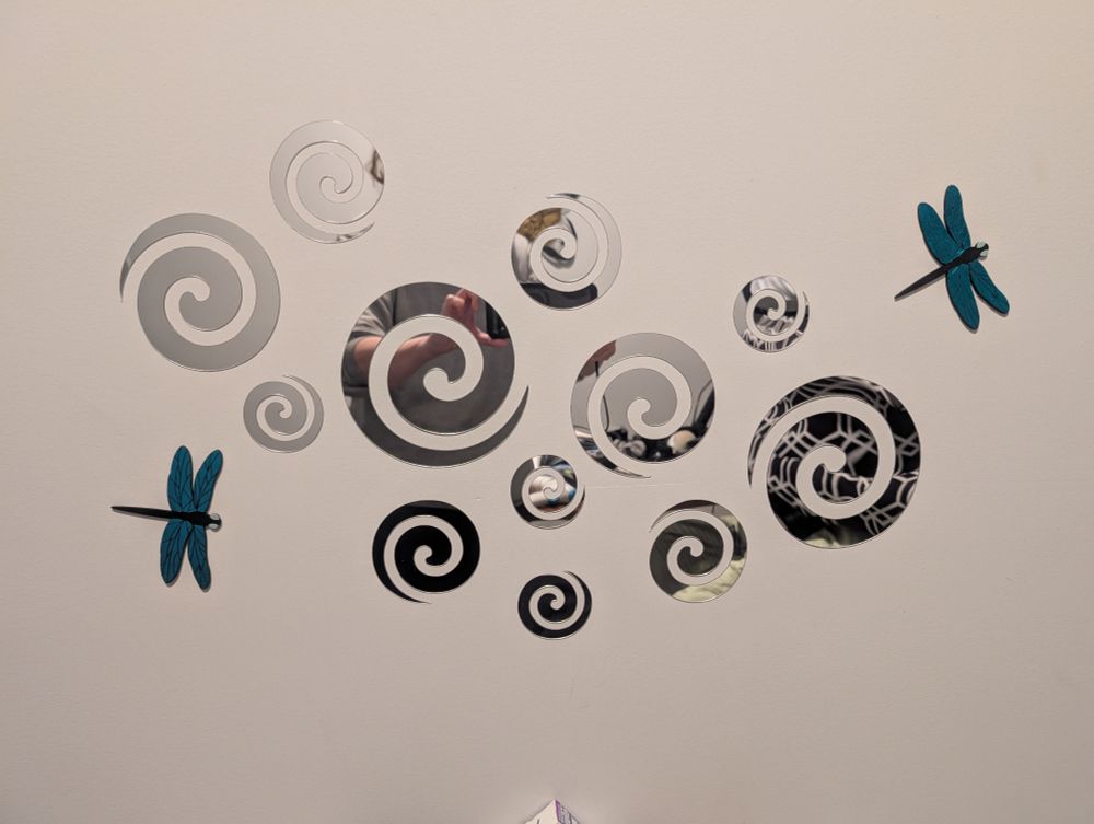 Mirrors cut into spiral shapes adorn a white wall, accented with two blue dragonflies. My phone and I are just barely visible in the spirals, and so is my black and white hexagon curtain.