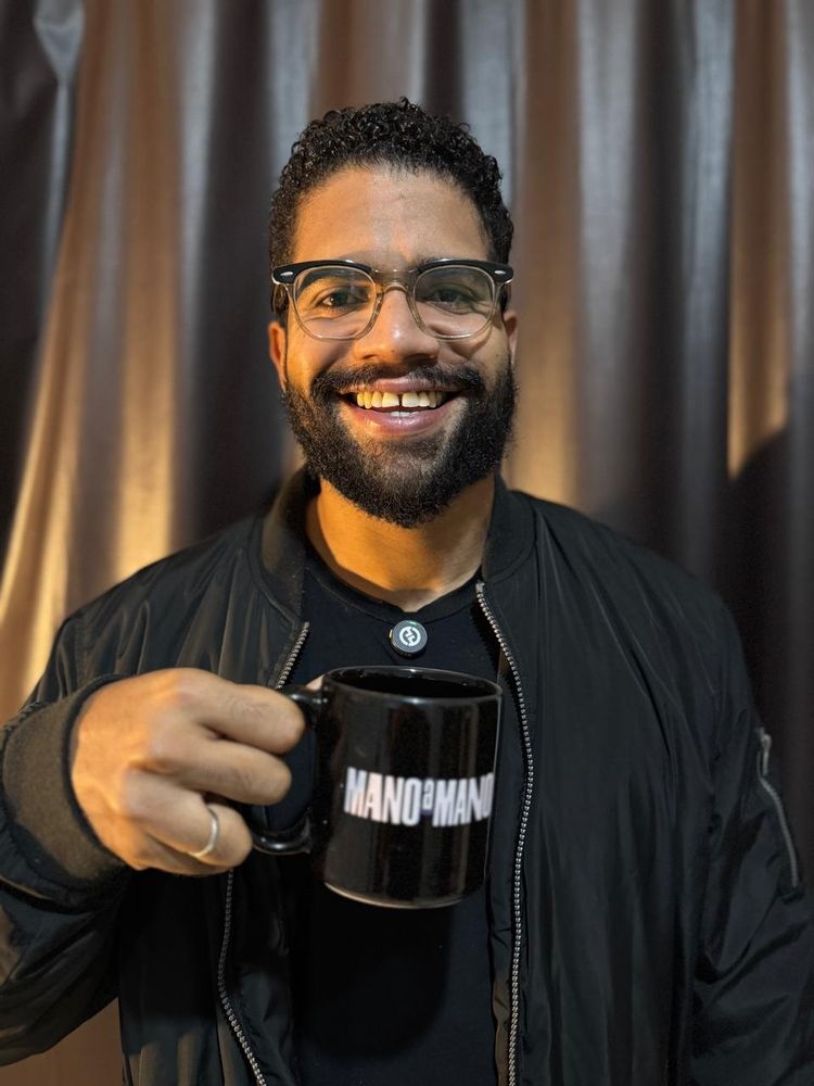 Jones Manoel sorrindo segurando uma caneca escrita "mano a mano". Ao fundo temos uma cortina prateada de estúdio de gravação.