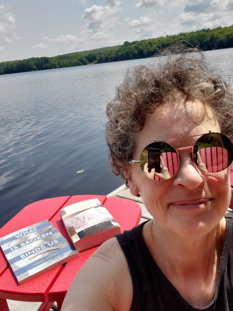 Me, mirror-sunglassed and smiling, on the dock at Big Doe Lake, with lake, far shore and fluffy-clouded sky behind me, with these books on a small red table behind me: What Is Broken Binds Us by Lorne Daniels (University of Calgary Press) and In Search of Lost Time Volume 1 Swann's Way by Marcel Proust (The Modern Library Classics)