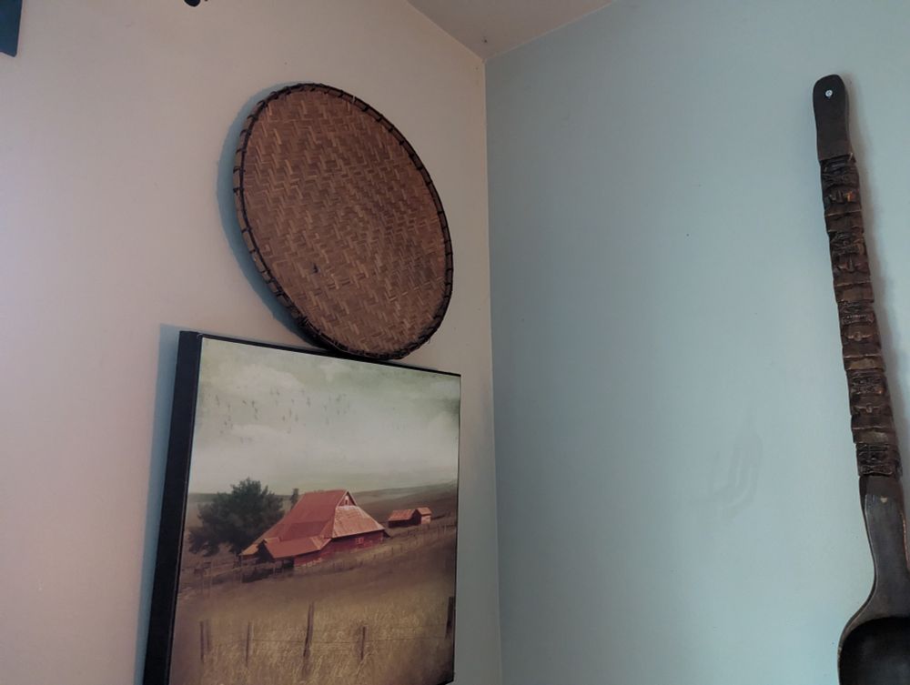A corner in my dining room.  A circular woven winnowing basket sits on top of a framed print of a red barn in a western plains landscape. 
