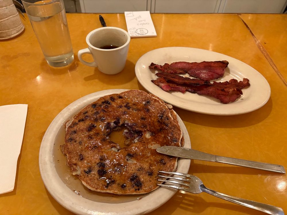 2 blueberry pancakes, 3 slices of bacon and a cup of coffee on a yellow Formica top