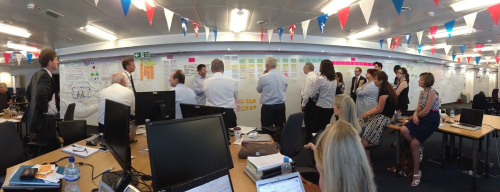 Photo of governance meeting during 2013 reset of Universal Credit. Photo includes lots of people standing up in front of a whiteboard wall containing many index cards. There is bunting.
