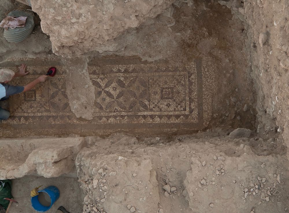 Photograph looking directly down on an archaeologist gently brushing a Roman mosaic which is long and narrow and features a pattern design