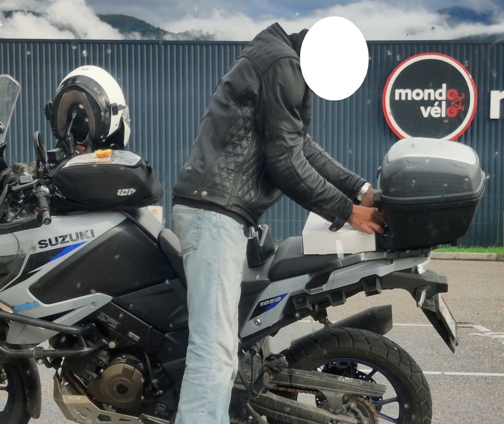 Motard occupé à scotcher une boite de gâteaux à l'arrière de la selle de sa moto en essayant visiblement d'être très économe sur le scotch.