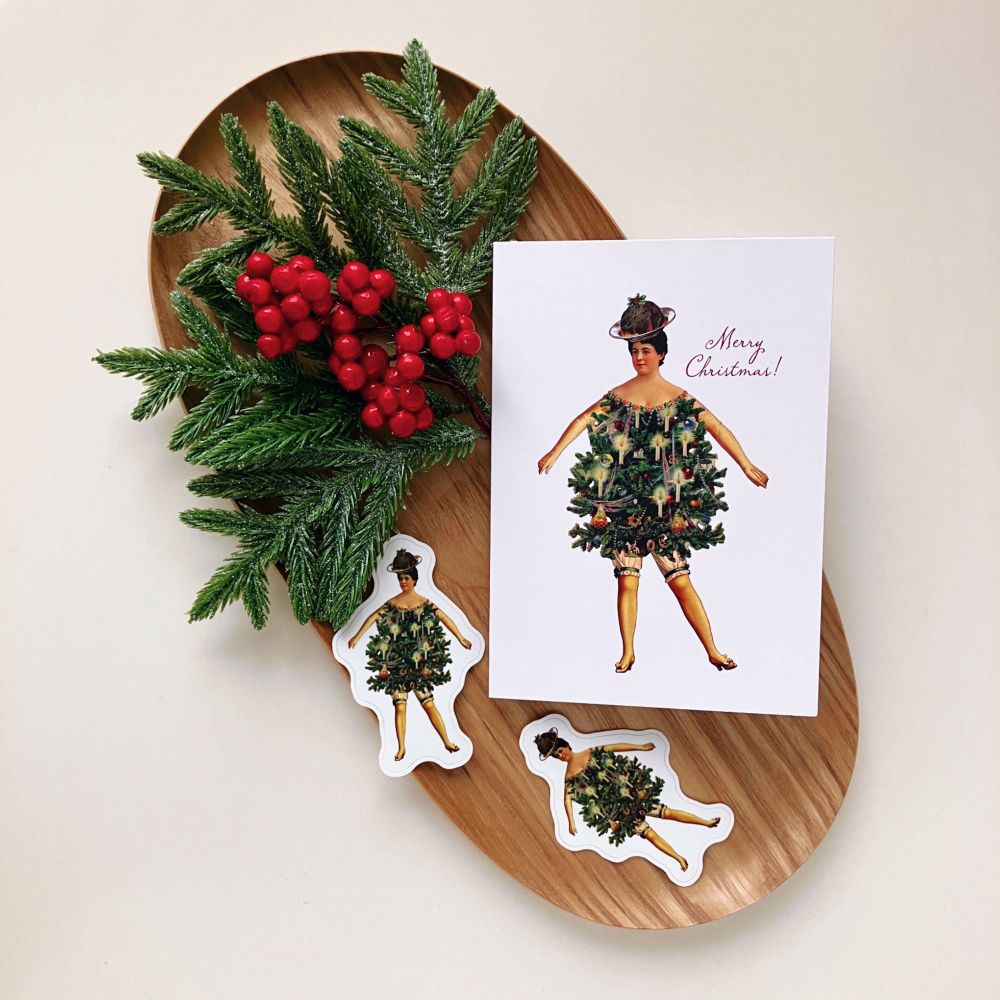 A card and matching stickers depicting a woman dressed as a Christmas tree, with a plum pudding on a dish worn as a hat. The card and sticker are on a wood tray decorated with a branch of evergreen and red berries