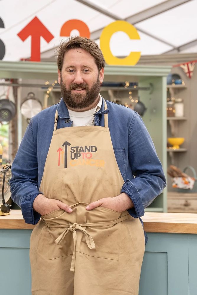 Promotional photograph of actor and writer Jim Howick inside the Bake Off tent. He wears a beige Stand Up To Cancer apron.