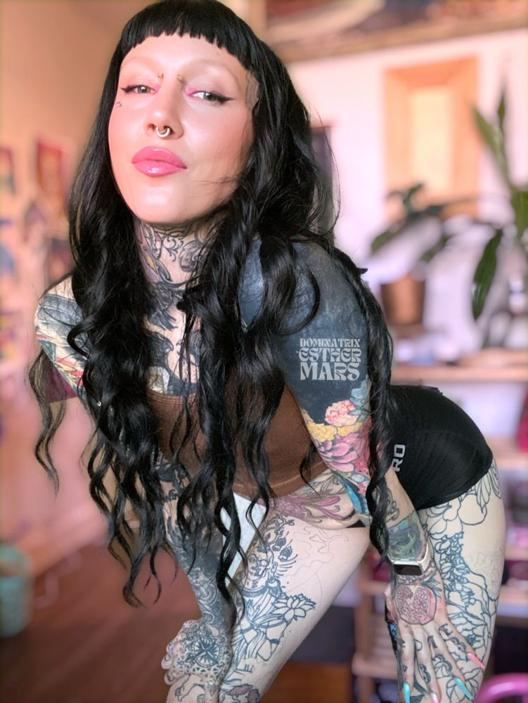 A non-binary femme with brown top, black shorts, black hair with short bangs, many tattoos, piercings bent forward with a smile and soft pink lip gloss. There is blurred out art and plants in the background.