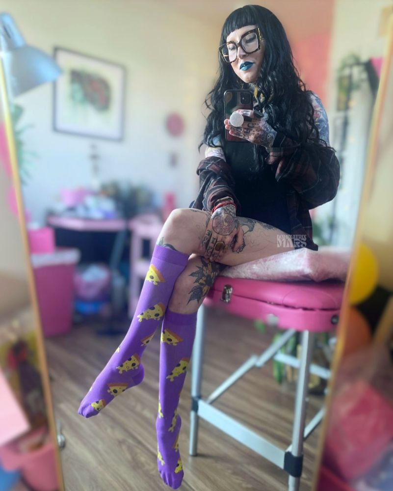 Person with long black hair and dark lipstick sits on a pink massage table, posing in front of a mirror. They wear purple knee-high socks with yellow cartoon pizza slices, black clothing, and oversized glasses. Their body is heavily tattooed, and the room is decorated in soft pink tones with plants and artwork in the background.