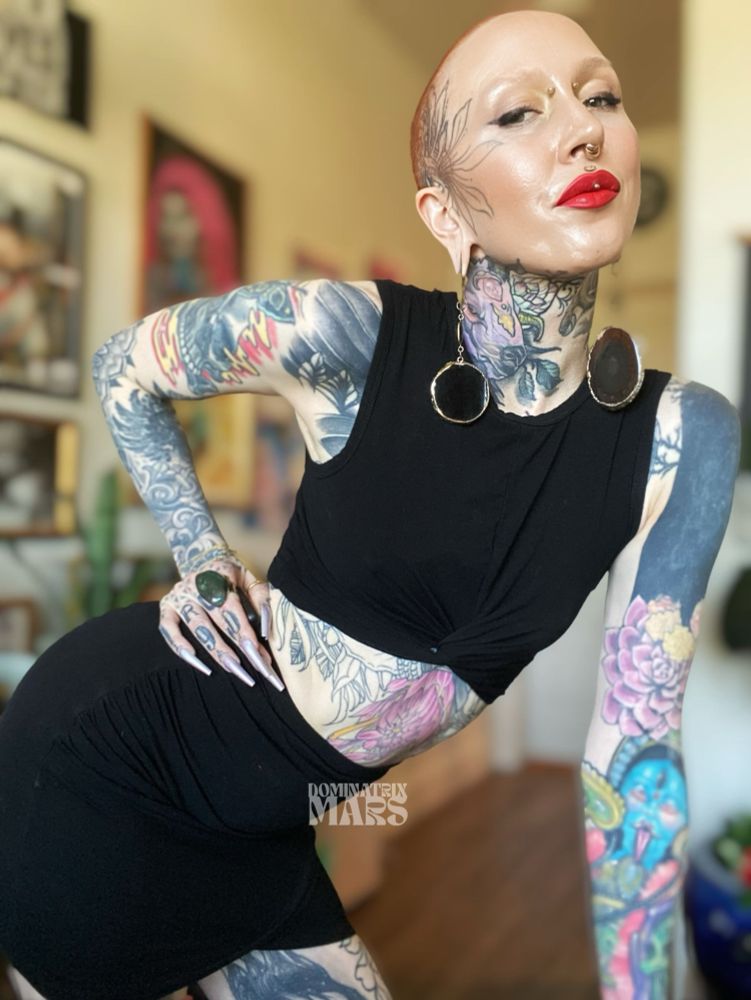 a non/binary femme figure with short buzzed red hair, a black crop tank, black skirt, blah and gold agate ear hangers, bloodstone ring, extra long chrome fingernails, facial piercings, and many tattoos. they are leaned forward with one hand on hip and one hand on surface. there is blurred out artwork and hardwood floors in the background. small watermark says "DOMINATRIX MARS"