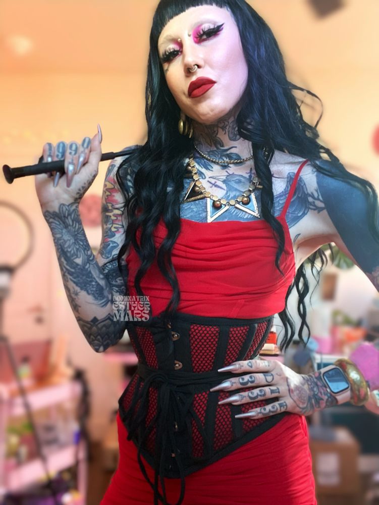 tattooed and pierced femme with long black hair, short bangs,  a red dress, black corset, long nails and holding a riding crop. 