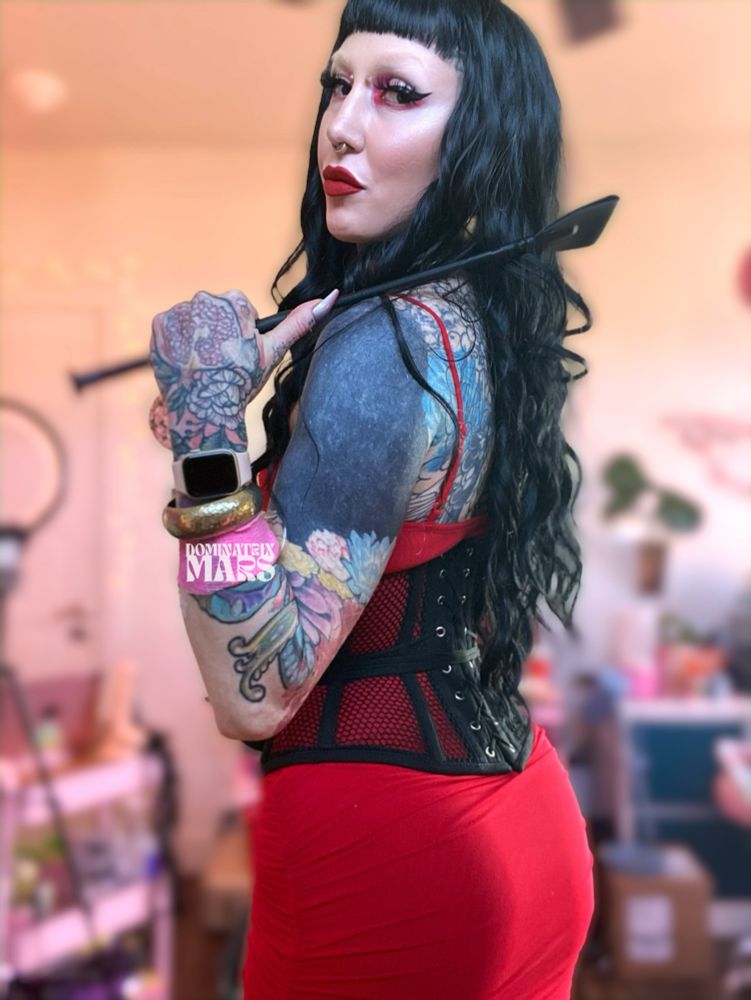 A non-binarry femme who is heavily tattooed and pierced. They have long curly black hair with short bangs, a red dress, and black corsette. They are holding a riding crop and have long fingernails. Dominatrix Sev Mars has glamorous eye makeup on with pink glitter inner color and long fake eyelashes and deep red lipstick.

The background is blurred out with plants, pink, and furniture in background. 