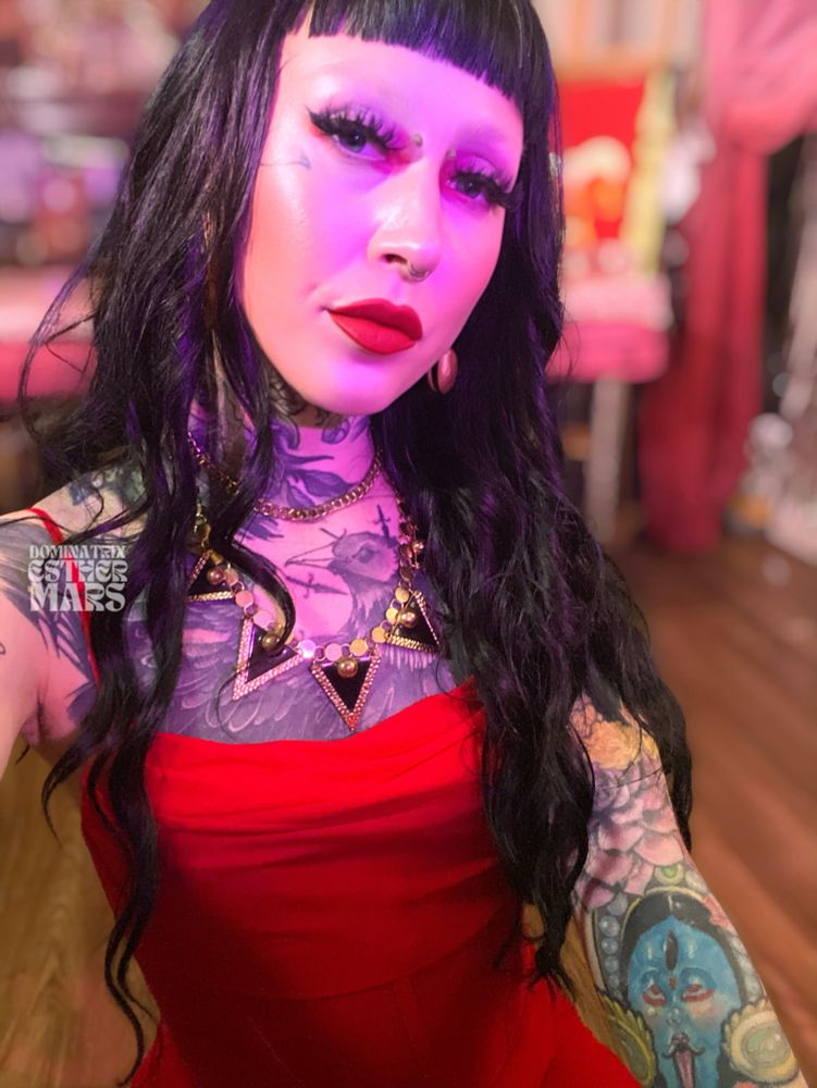 Alt text: A femme-presenting person with long wavy black hair and blunt bangs poses for a selfie in a warmly lit room with pink and red tones. They wear a bold red dress and dramatic makeup with bright red lipstick and long false lashes. Their skin is adorned with extensive tattoos, including a large black-and-grey chest piece of a bird and colorful designs on their arms. They wear a triangular statement necklace with gold accents and teardrop weighted brass earrings. The lighting casts a soft pink and purple glow over their face and surroundings. The watermark reads “DOMINATRIX ESTHER MARS.”
