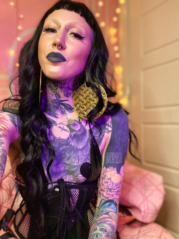 tattooed non-binary femme human with large woven mixed metal weighted jewelry, facial piercings, short bangs and long black hair in latex, heart pasties, corset against a soft pink background with string lights
