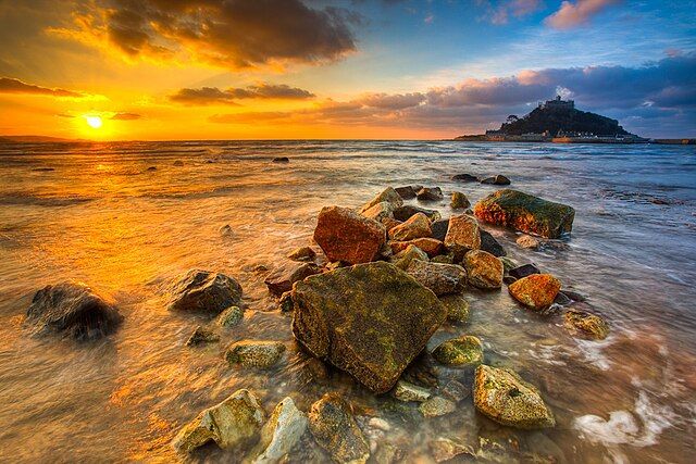 Colour photo of a sunrise over rocks and waves. This image is on Wikimedia Commons, with the following title: "St_Michael_Mount_6692_6697". If you have taken the time to access this text, may today be nothing but kind to you and yours.