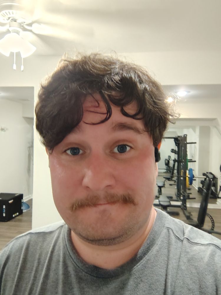 Sweaty white guy in a gym with brown hair, blue eyes, and a mustache in a gray shirt