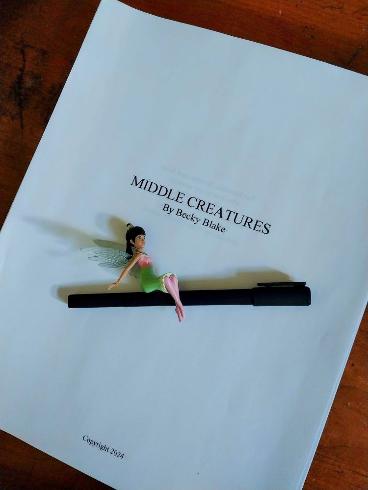 A pen sits on a printed out manuscript called Middle Creatures. A small fairy rides the pen like a broomstick.