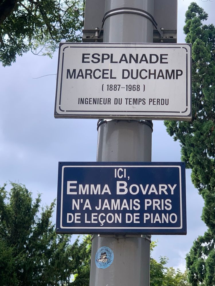Photo d’un poteau de lampadaire avec 2 panneaux de rue. 
En haut, le panneau est beige et le texte marron. Il est indiqué : ESPLANADE
MARCEL DUCHAMP (1887-1968)
INGÉNIEUR DU TEMPS PERDU
Le panneau du bas est bleu marine avec le texte blanc. Il est écrit : ICI, EMMA BOVARY N'A JAMAIS PRIS DE LEÇON DE PIANO. 
Pour des raison de cadrage, seul le panneau du bas est visible sur la photo de couverture. 