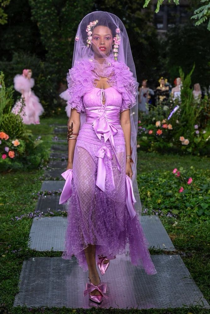 Model on runway wearing rich purple gown with mesh details satin bows