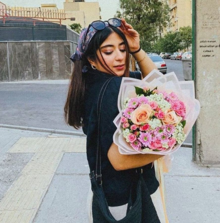 Photo de Parnia Abbassi. Elle est dans la rue, à moitié de dos, elle se tourne vers la caméra en relevant ses lunettes de soleil sur sa tête. Elle regarde un grand bouquet de fleurs roses qu'elle tient de l'autre main.
