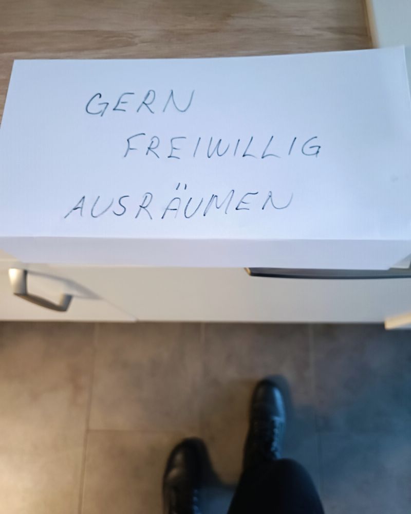 Ein handgeschriebenes Blatt mit dem Text "GERN FREIWILLIG AUSRÄUMEN" liegt auf einer Küchenanrichte, darunter ist die Spülmaschine. Meine Beine in schwarzen Hosen und schwarzen Boots sind auch noch unscharf am unteren Bildrand zu sehen