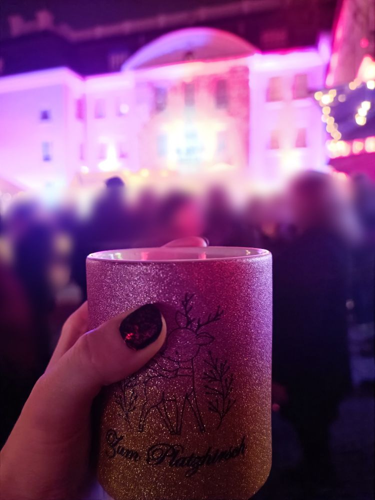 Ich halte eine gelb-rosa glitzernde große Tasse hoch, auf der ein kleiner Hirsch drauf ist, darunter steht "Zum Platzhirsch". Im Hintergrund ein gut besuchter Weihnachtsmarkt vor einem Schloss