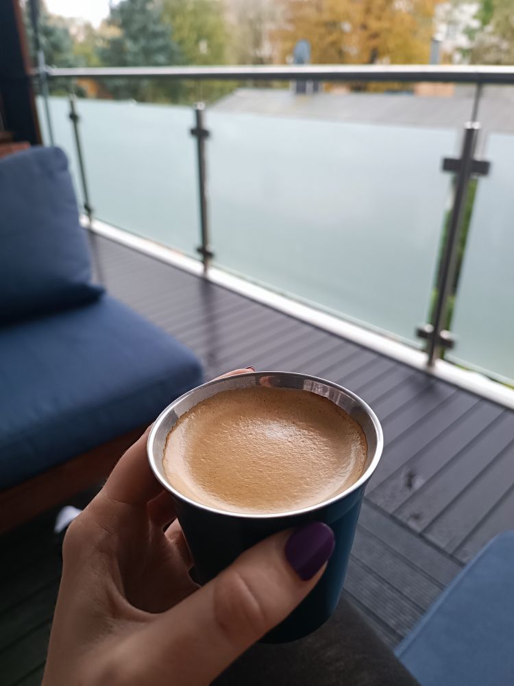 Meine Hand hält eine dunkelgrüne Metalltasse mit Kaffee und schöner Crema hoch. Ich sitze auf einem Balkon mit nassem Boden, im Hintergrund herbstlich gefärbte Bäume. Auf dem Balkon steht eine Sitzgruppe mit dunkelblauen Kissen