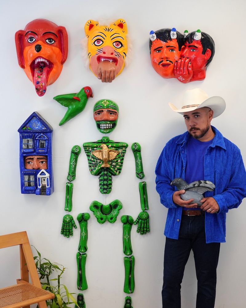 An artist stands next to a vibrant display ofcolorful papier-mâché sculptures on a white wall. The artwork includes a green skeleton, a parrot, masks depicting various emotions, and a blue house with a human face. The artist, dressed in a blue corduroy shirt, jeans, and a white cowboy hat, holds a gray pigeon sculpture while gazing thoughtfully.