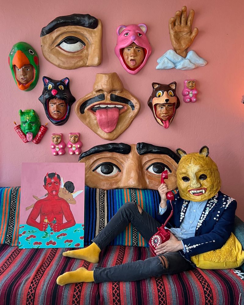 A person wearing a yellow animal mask and a blue jacket with pearl-like embellishments sits on a colorful striped couch holding a red rotary phone. Their face is obscured by the mask. Behind them, a pink wall is adorned with papier-mâché masks and sculptures, including large facial features, animal-themed headpieces, and small figurines. A painting of a red-skinned figure with horns a devil, holding two men, sits on the couch beside them. The scene is vibrant, playful, and surreal, with folk-art influences.