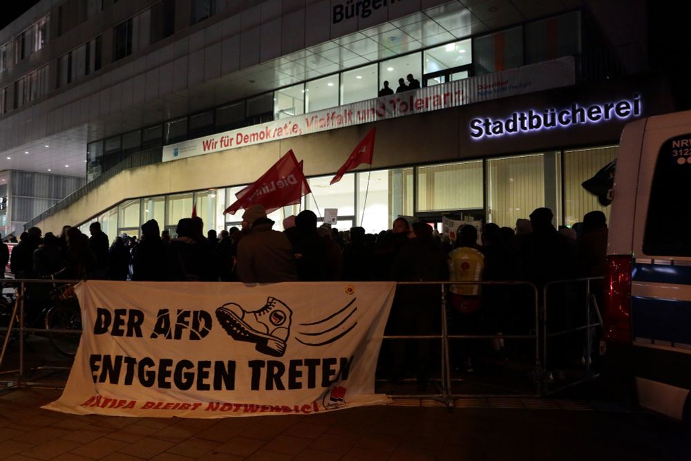 Blick auf den Gegenprotest. Im Vordergrund ein Banner "Der AfD entgegentreten- Antifa bleibt notwendig". Im Hintergrund das Bürgerhaus mit dem erleuchteten Veranstaltungssaal im ersten Stock, vor dem Polizei postiert ist.