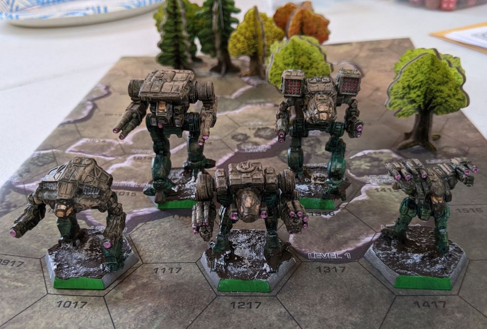 A clan star of battlemechs on a paper with a hex grid. Little cardboard trees are in the background. The mechs are brown and tan from the waist up, with dark green legs. Their cockpit windows are golden bronze, like a nasa helmet. The mud on their base is speckled with snow.
