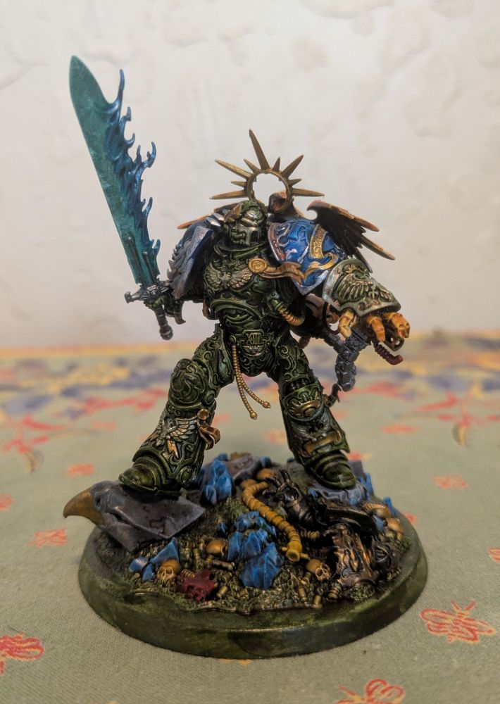 A model of Roboute Guilliman, with helmet on, painted in the color scheme of my own space marine chapter. The armor is green, the shoulders are blue, the backpack is yellow, the sword is cyan, and the trim is a mix of gold and silver.