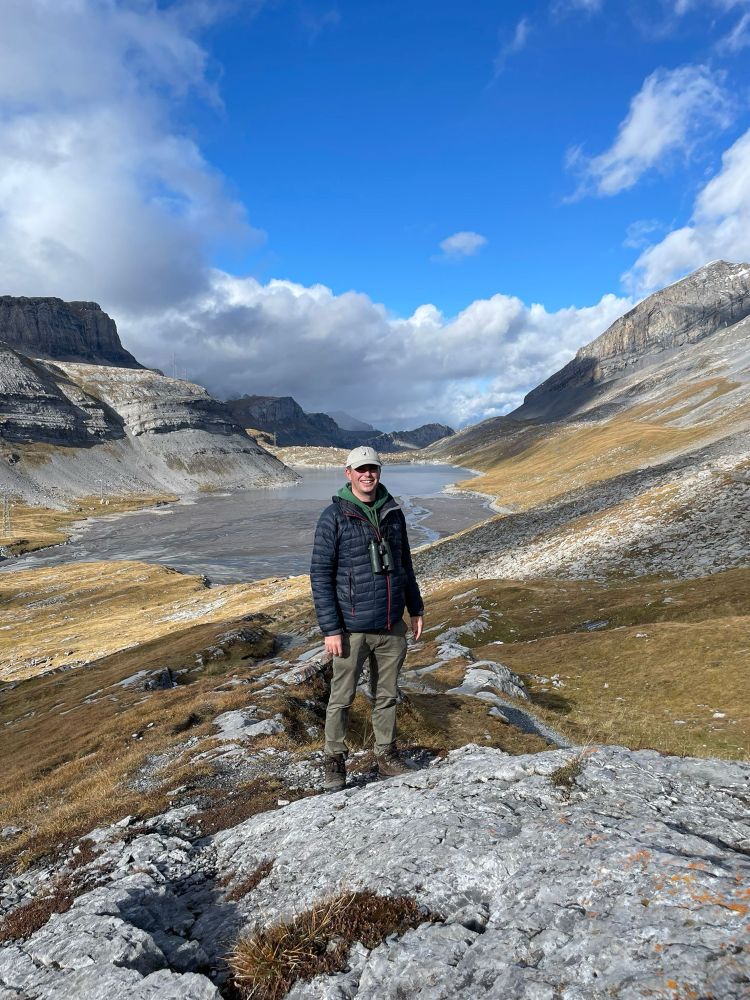 A photograph of me stood at Gemmi Pass wearing walking boats, green trousers, a navy windbreaker with a green hoody and a beige hat. The view behind me looking north includes the alpine pass with a lake in the mid distance. The slopes are largely rocky with some brown grasses. The sky is blue with some light grey cumulus clouds.