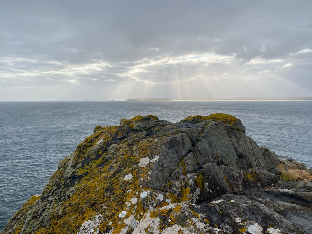 The view looking east from the lookout am below the NCI station over St Ives bay to Godrevy. In the foreground is a grainy rock covered in orange and white lichen. Beyond the rocks, the water of the bay is dark grey and beams of sunlight are filtering through grey clouds over the coast at Godrevy. 